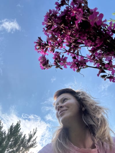 The image is an outdoor, low-angle shot of a woman looking up at a bunch of pink flowers against a blue sky with scattered white clouds. She has light brown, windblown hair, freckles, and is wearing a pink shirt. The flowers appear to be wildflowers. In the lower left corner, a dark green evergreen tree is visible. The sun is shining, creating a lens flare effect in the upper right portion of the image. The scene appears to be a natural outdoor setting, possibly a park or a garden. There is no visible indication of the specific location in the city of Чумна, Russia.
