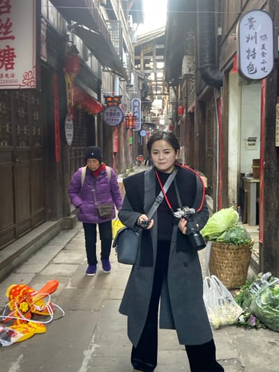 A woman with a camera poses in a narrow, old-fashioned street lined with wooden buildings adorned with signs and red lanterns. Behind her, an older person in a purple coat walks on the cobblestone street. A basket of produce sits near the woman, and a dragon-shaped decoration rests on the ground. The scene suggests a tourist spot or a traditional market.