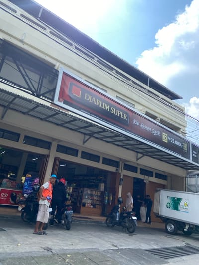 The image shows a storefront in Cimahi, Indonesia, located at latitude -6.872 and longitude 107.541. The store is a small shop with a large sign above the entrance that reads "Djarum Super" in red letters and "#Ini Rasanya Super" in white lettering. There are two men and a woman in front of the store. The man on the left is wearing an orange vest and a blue hat. The woman is wearing a headscarf and a long dress. The other man is wearing a black shirt and a blue hat. There are three motorcycles parked in front of the store, and a white delivery truck is parked on the right side of the image. The storefront is a small commercial building with a light grey exterior and white lettering. The building has a large awning over the sidewalk, and there is a white metal railing above the awning. The awning is covered with a white canopy, and there are several wires running across the awning. There are many signs and posters displayed on the exterior of the building. There is a clear blue sky with some fluffy white clouds in the background. The street is made of concrete. The image was taken during the day, as there is a lot of light in the image. The image is well-lit, and the scene appears to be clear and sharp. The people in the scene are stationary and seem to be engaging in a transaction at the shop.  The delivery truck is parked directly in front of the building, likely dropping off goods. The image is a typical street scene in Cimahi, Indonesia.