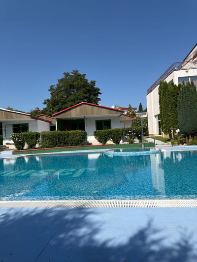 The image depicts a resort with a swimming pool in the foreground and buildings in the background. The pool is filled with clear blue water and surrounded by a blue tiled deck. The buildings appear to be bungalows with white walls and red-tiled roofs, situated behind a manicured hedge. To the right, a larger white building with balconies is visible, surrounded by tall green trees. The sky is clear and blue, suggesting a sunny day. The scene is peaceful and serene, typical of a vacation destination. Based on the visual cues, this location could be in Byala, Bulgaria, a coastal town known for its resorts and beaches.
