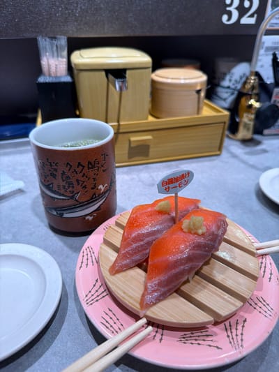 Two pieces of salmon nigiri adorn a wooden platform atop a pink patterned plate, accompanied by chopsticks. Beside it, a brown mug featuring Japanese script and a fish illustration sits near a small white plate. A beige bamboo-style container and a black menu with the number "32" are visible in the background. The table appears to be light gray.