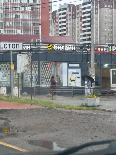 In Novoe Devyatkino, Russia, an outdoor street scene depicts a row of shops under a cloudy sky. A prominent sign with "СТОП" (STOP) is visible on the left, next to a sign for "БИЛАЙН" (BEELINE) with its distinctive yellow and black logo. Further to the right, a sign reads "БАР Р" (BAR R) with colorful bottle graphics. The shops have large glass windows with decorative metal grilles. Two individuals are present: one person in a brown coat walks past the storefront, and another person with an umbrella walks by a planter with orange flowers in front of a shop. The street in the foreground is wet and appears to have puddles, suggesting recent rain. Tall apartment buildings form the background.