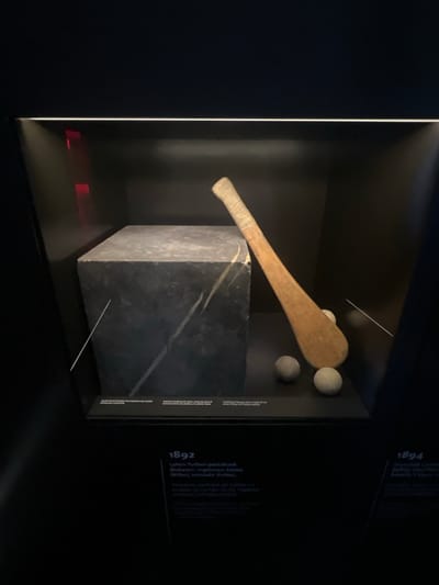 The image depicts a museum display case containing artifacts related to Basque sports. Inside the case, a wooden bat-like implement and three round balls are arranged next to a large, dark, rectangular stone block with lighter veining. The display is illuminated from above, casting shadows. Below the display case, text is visible on a dark background. On the left, text labeled "1892" is in white, followed by smaller white text in Basque and Spanish. On the right, text labeled "1894" is also in white, with accompanying smaller text. The setting appears to be an indoor exhibition space. The text suggests the display is located in Bilbao, Spain, as it references Basque sports and the year 1892.