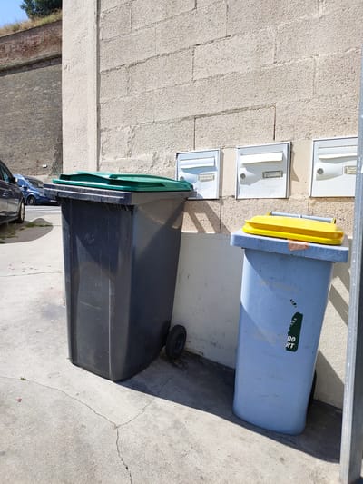 This image depicts a scene outdoors, likely on a sunny day, with a textured, light-colored brick wall forming the background. In the foreground, two large garbage bins are prominent. The bin on the left is dark gray with a green lid, and it has wheels on the bottom. To its right is a light blue bin with a bright yellow lid. A sticker with text is visible on the side of the blue bin, partially obscured, but "00 RT" is legible. Above the bins, mounted on the wall, are three white mailboxes with their flaps down. The mailbox on the far right has some writing visible, possibly a name or address.
The ground is concrete, with visible cracks and some sparse green shoots. To the left of the garbage bins, a portion of a dark-colored car is visible, suggesting a parking area or street. In the upper left corner of the image, a glimpse of a brick wall, possibly a historical fortification or embankment, topped with dry grass, is visible, hinting at the broader location. The sunlight casts distinct shadows, indicating it is likely midday. The overall impression is one of an everyday urban or semi-urban setting. No people are visible in the frame. The mood is neutral and observational.