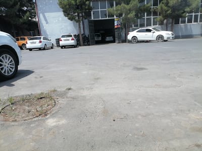 The image shows a street scene in Juma, Uzbekistan, featuring several cars and a building that appears to be a service station or garage. A white sedan, possibly a Chevrolet Malibu, is parked on the right side of the street, facing left. Another white sedan, a Chevrolet Lacetti, is parked in front of it, also facing left. In the background, an orange hatchback, likely a Daewoo Matiz, is parked. The building in the background has large windows and an open garage door, with two white vehicles inside. Piles of tires are stacked to the left of the garage entrance. The ground is paved with asphalt, showing some wear and tear, including a circular metal manhole cover with grass growing around it. Pine needles are scattered on the pavement. The lighting suggests it is daytime, likely late morning or early afternoon, with bright sunlight casting shadows.