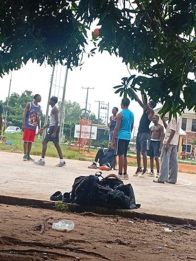 This media file depicts a group of young men on an outdoor basketball court in Ukukwa, Nigeria. The scene is framed by lush green foliage from trees, suggesting it's likely daytime, possibly overcast given the diffuse lighting. Several individuals are actively engaged in what appears to be a casual basketball game or practice.
In the foreground, a large black duffel bag rests on the dirt beside a basketball court. The court itself is a concrete surface with visible markings. Two young men are prominently featured in the center, one wearing a blue and purple basketball jersey with red shorts, and the other in a grey tank top and dark shorts. They are facing each other, possibly interacting during a pause in the game.
Further back, other individuals are visible. One man in a black tank top and shorts is reaching up towards a tree branch. Another person, shirtless with dark shorts, stands nearby. A man in a light-colored, patterned shirt and grey pants is also present on the court. On the left side of the frame, a man in a dark shirt is seated on the ground, and another man in a blue tank top and shorts stands observing.
In the background, a white car is parked, and a building with a sign that reads "SCHOOL OF GENERAL STUDIES" is visible. Utility poles and other structures suggest an urban or semi-urban environment. The overall mood appears relaxed and focused on the sporting activity. There are no overt displays of strong emotion, but the engagement in the game suggests camaraderie and active participation. The presence of the school sign provides a specific location cue within Ukukwa, Nigeria.