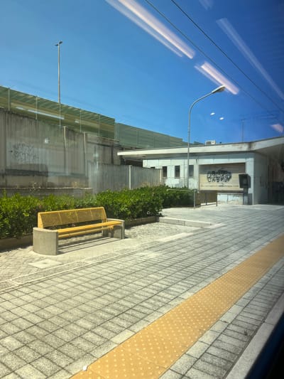 The image depicts a scene at a public transportation stop, likely a train station or bus stop, in Bari, Italy. In the foreground, a yellow slatted bench is situated on a paved area with a textured yellow tactile paving strip running parallel to what appears to be a platform edge. The paving is composed of square gray tiles. Behind the bench, a dense green hedge borders a concrete wall adorned with graffiti. Further back, a low-rise building with a rolling shuttered entrance and a few windows is visible. A streetlamp stands to the left of the building, and another is seen in the distance. The sky is clear and blue. Reflections of fluorescent lights are visible in the upper portion of the image, suggesting the photo was taken from inside a vehicle or through a window. The overall impression is one of an urban public space on a sunny day.