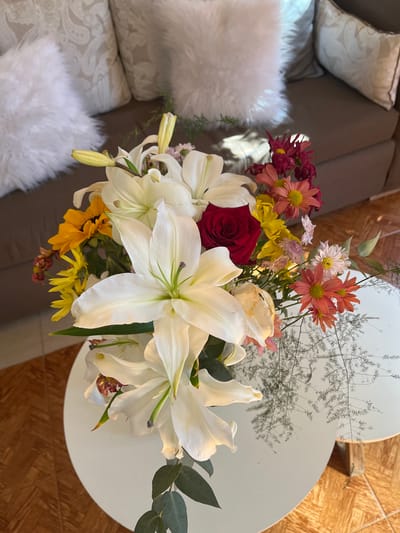 The image shows a floral arrangement on a white, two-tiered, oval-shaped table. The arrangement features large white lilies, a red rose, and various smaller flowers in shades of orange, yellow, and pink. Some greenery is also present. The table is situated on a wooden floor with a parquet pattern. In the background, part of a gray couch is visible, adorned with light-colored pillows. The location is San Fernando del Valle de Catamarca, Argentina.