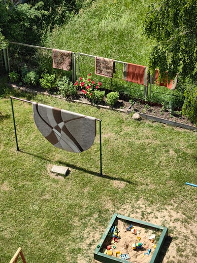 The image captures an outdoor scene, likely a backyard in Myadzyel, Belarus. A metal fence runs horizontally across the upper portion of the frame, adorned with four colorful towels or small rugs hanging to dry. Behind the fence, dense green vegetation, including trees and tall grass, creates a natural backdrop.
In the foreground, a large, semi-circular rug with a geometric pattern is draped over a green metal drying rack. The rug is suspended in mid-air, casting a shadow on the lush green grass below. A flat, grey stone rests on the grass near the drying rack.
To the lower right of the image, a square sandbox is visible, filled with sand and scattered with various children's toys, including small trucks, a cement mixer, and digging tools. The sandbox appears to be green-trimmed. The ground surrounding the sandbox is a mix of grass and bare earth. A sliver of a blue plastic hose can be seen on the far right edge of the frame. The overall impression is one of a sunny day with domestic activities taking place in a suburban or rural setting.