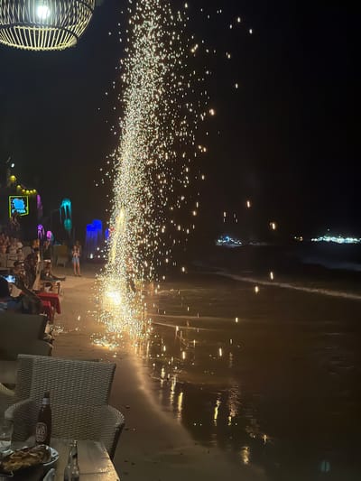 The image depicts a nighttime scene on a beach in Ao Phai, Thailand. A large, ground-based pyrotechnic device, emitting a dense vertical spray of golden sparks, is active near the waterline in the mid-ground, illuminating the wet sand and casting reflections. To the left of the pyrotechnic display, multiple individuals are gathered on the sand. Some are seated at low tables with chairs, while others stand observing the display. In the immediate foreground, an overhead wicker lantern with a single bulb is visible in the upper left corner, casting light downward. Below it, a table holds a brown bottle identified as "LEO" beer, another glass bottle, and a plate containing food items resembling chips or nachos. A wicker chair is positioned to the right of this table.
Further into the background on the beach, a series of illuminated decorative structures resembling glowing jellyfish or fantasy plants in blue, pink, green, and yellow hues are visible. Other groups of people are scattered across the beach area. In the far background, across the water, distant clusters of artificial lights indicate a shoreline or vessels. The ocean shoreline shows gentle waves breaking, with light reflecting off the wet sand and water. The overall environment suggests an evening gathering or event at a beachside establishment.