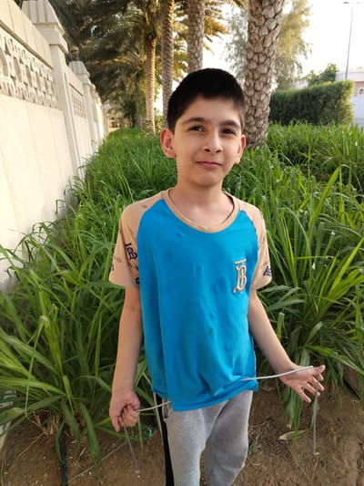 A young boy stands in front of a green, grassy area with palm trees in the background. He is wearing a blue t-shirt with beige sleeves and gray sweatpants. The boy is holding a thin, metal wire in his hands. He appears to be outdoors, possibly in a park or garden, given the presence of lush vegetation and palm trees. The lighting suggests it is either early morning or late afternoon, as the sun casts a warm glow on the scene. The boy's expression is neutral, and he looks directly at the camera. The background includes a white, decorative wall, and further beyond, more greenery and parts of buildings. The overall scene is peaceful and suggests a casual outdoor setting in Al Mas'oudi, United Arab Emirates. There is visible text on the sleeves of the t-shirt, featuring a repeating pattern of "tb" and a stylized "B" logo.