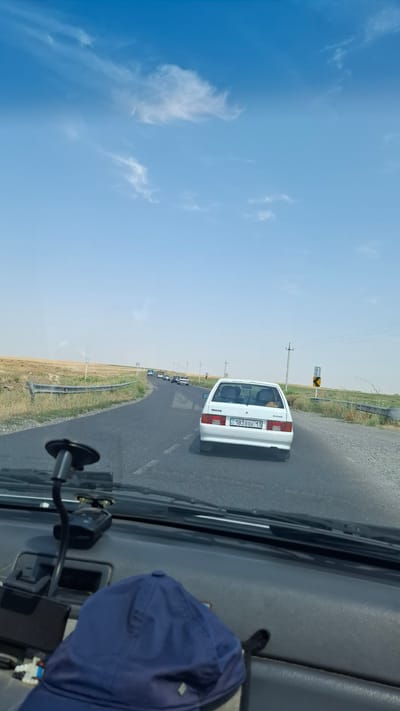 This image depicts a view from inside a vehicle driving on a road, likely in or near Теспе, Kazakhstan, under a bright, clear sky with scattered clouds. The road appears to be a two-lane highway with a guardrail on the left side and a dry, grassy landscape on either side. Several cars are visible on the road ahead, traveling in the same direction. A white sedan is prominently featured in the foreground on the right side of the road, displaying a license plate that reads "C 183BBK 13". The dashboard of the vehicle from which the photo is taken is visible in the foreground, with a blue cap and a dashcam mounted on the windshield. Utility poles are present along the roadside. The overall environment suggests a rural or semi-rural setting.