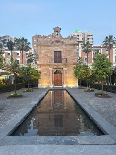 This is a vertical, eye-level, full shot of a central courtyard at dusk in Malaga, Spain, likely the courtyard of the Palacio Episcopal, featuring a long, narrow reflective pool. The pool is positioned in the foreground and reflects the facade of the historic building. The building, constructed of large sandstone blocks, has a large wooden door and a smaller door above it, both with ornate metalwork. A bell tower with a bell is visible above the central structure. The courtyard pavement is made of grey cobblestones. On either side of the pool are rows of young trees in planters. Behind the historic building, modern apartment buildings are visible, including one with the Unicaja logo. The sky is a pale blue, indicating twilight. There are no people visible in the immediate foreground or engaged in any specific actions. The scene is still and tranquil.