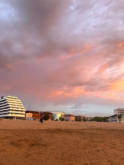 The image depicts a beach in Gijón, Spain, during sunset. The sky is filled with dramatic clouds painted in hues of pink, orange, and purple. In the foreground, a sandy beach stretches out, with the texture of the sand clearly visible. A young woman with purple hair, wearing a black dress and sneakers, is captured mid-action, seemingly jumping or dancing on the sand. She is positioned in the middle ground of the image.

Behind the beach, a cityscape emerges. On the left, a distinctively modern, multi-tiered white building with dark horizontal bands stands out. To its right, other buildings of varying architectural styles and heights are visible, including one with a glass facade reflecting the sunset colors. Trees and poles suggest urban infrastructure and greenery. The overall scene conveys a dynamic atmosphere, capturing a moment of freedom and beauty against the backdrop of a coastal city at dusk.