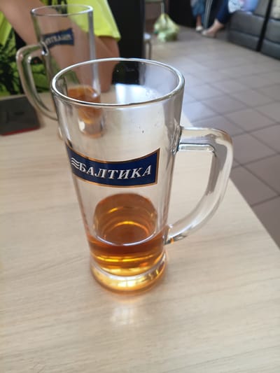 This media file shows two beer mugs on a light-colored wooden table. The mug in the foreground is tilted, revealing golden-colored liquid inside and a blue label with white text. The text on the label reads "БАЛТИКА". The background is blurred but shows what appears to be a tiled floor and a person sitting on a sofa in the distance. The lighting suggests it is daytime. The setting appears to be indoors, possibly a cafe or a home.