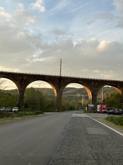 The image, rotated 90 degrees clockwise, depicts a grand stone viaduct with multiple arches, presumably a railway bridge, stretching across a scenic valley. The sky above is cloudy, with patches of sunlight creating a warm, golden glow, suggesting late afternoon or early evening. Lush green hills and trees are visible through the arches of the bridge, highlighting the natural landscape.

Parallel to the viaduct, on the right side of the frame, runs a paved road with a clear white line. Several vehicles are parked along this road, including what appears to be a red utility truck or fire engine, and a few cars. Utility poles or power lines are visible running along and above the viaduct, indicating active railway infrastructure.

A blue road sign with white text is prominently displayed on the right side of the road, showing the German words "Camping," "Gasthof" (guesthouse), and "Feuerwehr" (fire department). Below this, a standard circular speed limit sign indicates "80". The scene is devoid of people and conveys a sense of tranquility. The provided location context for this image is Usoika, Bulgaria.