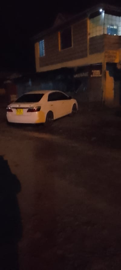 At night, a white sedan parks on an unpaved road in front of a two-story building with lit windows. The car's license plate is visible, though indistinct. Shadowy figures are present in the foreground. The lighting is dim, casting the scene in darkness. The overall atmosphere is subdued and quiet.