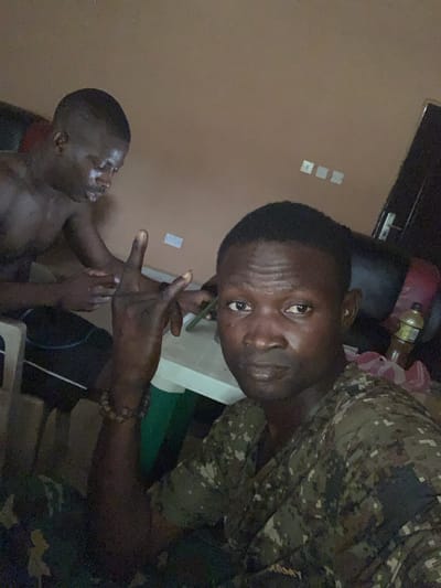 The image depicts two adult males within an indoor setting in Kado, Nigeria. In the immediate foreground, a male with dark skin and a short haircut, wearing a pixelated camouflage patterned shirt, is positioned looking directly at the camera. His right hand is raised, displaying a gesture with the index and pinky fingers extended, and a beaded bracelet is visible on his wrist. Behind and to the left, a second male, shirtless with dark skin and a short haircut, is seated on a white plastic chair, facing away from the camera and looking downwards, seemingly engaged with an object on a white table situated between the two individuals. The background consists of a plain light brown wall featuring multiple white electrical outlets or switches. A dark vertical element resembling a doorway is visible on the far right, where a clear plastic bottle is also partially observed. The room is dimly lit.