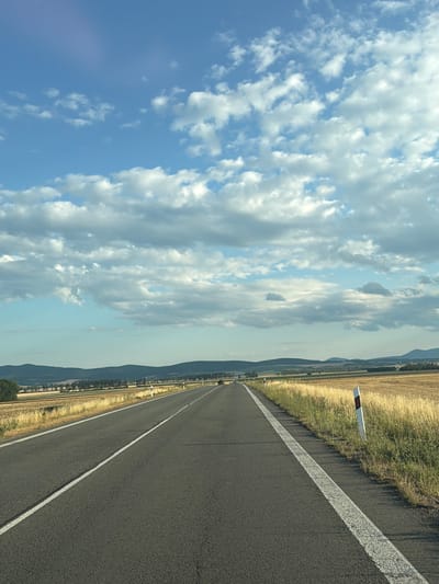 The image depicts a scenic view of a straight, asphalt road receding into the distance. The road is flanked by dry, golden fields on the right, with a line of trees and rolling hills in the background. The sky is a vibrant blue, adorned with scattered white cumulus clouds. The scene appears to be set in a rural area outside the city of Trebišov, Slovakia, likely during the late summer or early autumn harvest season. There are no people or vehicles visible on the road or in the immediate vicinity. The overall impression is one of tranquility and open space.
