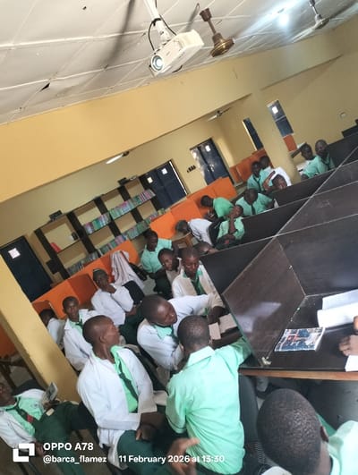 The image captures an indoor scene within an educational facility in the city of Rinze, Nigeria. Approximately twenty-five young males, primarily dressed in light green short-sleeved shirts, dark green ties, dark green trousers, and white lab coats, populate the room.
The environment features light yellow walls and a white ceiling composed of tiles, some exhibiting minor damage. Illumination is provided by recessed fluorescent lights and two brown ceiling fans. A white projector is mounted on the ceiling, directed away from the camera. The room is furnished with dark brown partitioned desks or cubicles where students are seated. In the background, against the left wall, shelves filled with books are visible, alongside orange-colored dividers. Two dark blue doors are present in the background, with one partially open, and barred windows are also visible.
Students are engaged in various activities. Several individuals are seated at desks, reviewing books or papers. Others are interacting in small groups, appearing to discuss or collaborate. Books and documents are present on multiple desks. A laptop is visible on a desk in the far background on the left side. One student in the lower left corner is leaning back, holding a small book or pamphlet.