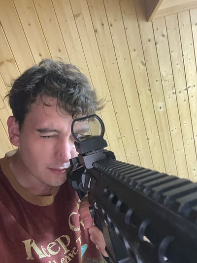A young man with wavy brown hair, wearing a maroon t-shirt with yellow trim, is aiming a black assault rifle with a red dot sight. He is looking through the sight with his left eye, his right eye is closed. The background is a wooden wall with vertical planks. The scene is indoors, in a room with a sloped ceiling made of the same wood. There are no discernible features that place the image in Vasilyevo, Russia.