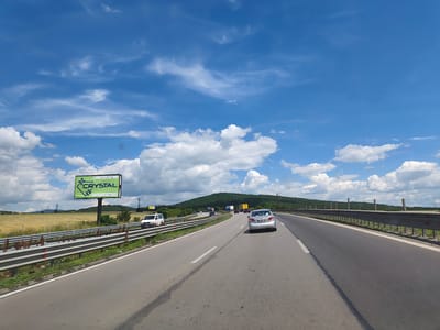 The image depicts a sunny day on a highway in Eleshnitsa, Bulgaria. A silver car is traveling on the right lane of the highway, with a guardrail on the right side of the road and a field of wheat on the left. In the distance, a green billboard for "CRYSTAL" is visible next to the highway. The sky is blue with scattered white clouds, and a green, forested hill is in the background. Several other vehicles, including trucks and a white van, are also present on the highway. The road appears to be well-maintained.
