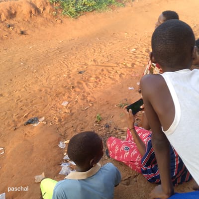 Four children sit on a sandy area littered with trash. One child holds a smartphone. The location is Nsugbe, Nigeria.