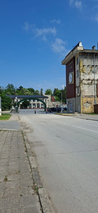 This is an outdoor street scene captured in Strazhitsa, Bulgaria, during bright daylight under a clear blue sky with scattered white clouds.

The image depicts a street with an asphalt road and a concrete sidewalk to its right. On the left side of the street stands a large, multi-story building with a weathered facade, showing signs of age with cracked plaster and exposed bricks. A prominent feature of this building is a large clock face centrally placed on a brown section. Above the clock, text is visible, reading "ВИНОПРОИЗВОДСТВО" (Winemaking/Wine Production) and below it, the year "1966". Further up on the weathered yellow wall of the same building, the year "1948" is painted.

Further down the street, on the left, another multi-story building with a more contemporary appearance is visible, featuring numerous windows. In front of this building, a distinctive green archway, possibly a decorative entrance or monument, spans part of the view.

Several people are present in the scene; a few individuals are standing or walking on the sidewalk near the green archway and further down the street. Several cars are parked or moving in the distance along the street on the right. Lush green trees are abundant, especially in the background and surrounding the buildings, indicating a green environment. A red "STOP" sign is partially visible on the right side of the street. The time of day appears to be midday or early afternoon given the bright lighting and minimal shadows.