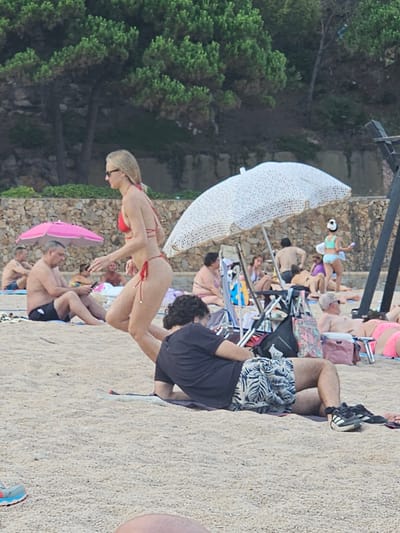 This image captures a beach scene in Blanes, Spain, with several individuals enjoying a sunny day. In the foreground, a person wearing a black t-shirt and patterned shorts is lying on the sand, with athletic shoes nearby. Behind them, a woman in a red bikini is walking, with a large white umbrella to her right. Further back, other beachgoers are visible, some sitting under pink and white umbrellas, and others relaxing on towels or chairs. The environment is sandy, with trees and a stone wall forming the backdrop, suggesting a coastal setting.