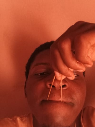 A close-up, front-facing shot shows a person's face from the nose down, with their hand holding a rubber band across their upper lip. The person's skin tone is dark, and they have dark, curly hair. The background is a solid orange color. The image appears to have been taken in an indoor setting, likely a home. The person's eyes are partially visible. The overall lighting is warm and orange-toned. There is no discernible information in the image that can be used to determine the location in the city of Bauchi, Nigeria.
