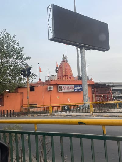 The image depicts a temple in Ahmedabad, India, likely during daylight hours under an overcast sky. The temple is an orange structure with traditional Indian temple architecture, including a domed roof adorned with decorative carvings and small spires. Orange flags are visible on the temple's roof.
In front of the temple, a large black digital billboard stands on a metal pole, tilted upwards. Adjacent to the temple, a yellow pole with a railing and a green metal fence runs along a paved area, possibly a roadside. A traffic light with three black signals is also visible, positioned near a leafy green tree.
A blue and white sign on the temple reads "Orbit" with smaller text below indicating "WIRES & CABLES". Another sign in a script that appears to be Gujarati is also visible on the temple wall. There are two air conditioning units mounted on the orange wall of the building. The overall scene suggests an urban environment with a mix of religious and commercial elements. No people are clearly visible in the image, but the presence of a traffic light implies an active area.