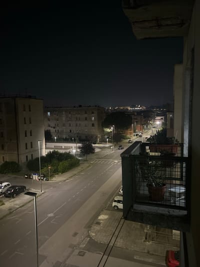 The image captures a nighttime view from a balcony overlooking a street in Benevento, Italy. The scene is characterized by artificial lighting from streetlamps and building windows, illuminating a residential area.
In the foreground, the balcony railing, made of dark metal, is visible. A large planter containing a potted plant sits on the balcony. The balcony itself appears to be part of an apartment building, with a concrete edge that shows some signs of wear.
Beyond the balcony, a two-lane street runs through the frame, bordered by sidewalks and buildings. Several parked cars are visible on the side of the road, including a black sedan, a white hatchback, and a red car further down. Streetlights line the road, casting pools of light.
The buildings are multi-story residential structures, appearing to be apartments. Some windows are illuminated, suggesting occupancy. In the distance, a cityscape with more buildings and lights can be seen, indicating a larger urban area. The sky is dark, typical of nighttime.
The overall impression is a quiet, urban night scene in Benevento.