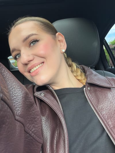 A young Caucasian woman with blonde hair in a braid and light-colored eyes is taking a selfie in the driver's seat of a car. She is wearing a maroon leather jacket over a black shirt and a small white quatrefoil earring in her left ear. Her makeup includes winged eyeliner and blush. The interior of the car, specifically a black leather headrest and seat, is visible. Outside the car window, greenery and a building can be seen. The car appears to be parked, possibly near a residential area, which is consistent with the location of Dreiliņi, Latvia, a suburban municipality known for its residential developments.