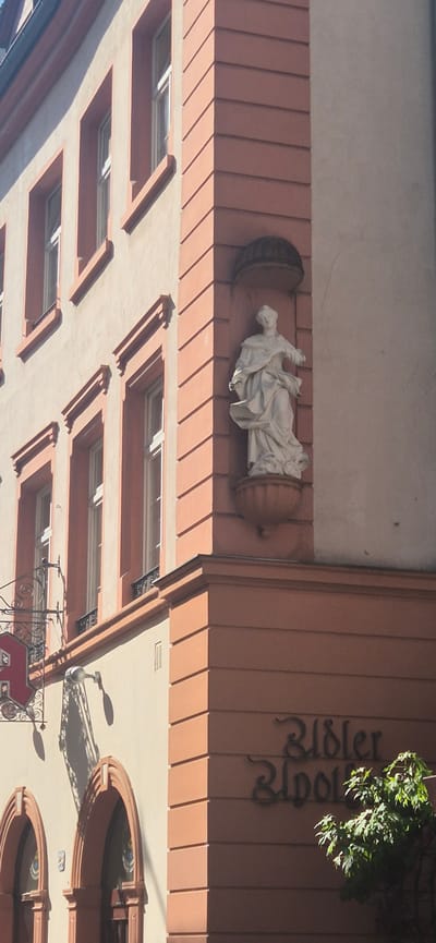The image shows a close-up view of the facade of a building in Mainz, Germany. The building has light beige walls with reddish-orange architectural details around the windows and along the edges of the building.
On the upper right side of the building, there is a white statue of a draped figure standing in a niche. Below the statue, on the reddish-orange wall, there is ornate dark lettering that reads "Riebler Apotheke." A green tree with lush foliage is partially visible in the lower right corner, obscuring some of the lettering.
Further to the left, on the beige section of the wall, there are several tall, narrow windows with white frames. A decorative iron signpost with a red emblem is attached to the building above a shop entrance. This entrance features arched openings with reddish-orange trim. The overall impression is of an older, well-maintained building in an urban setting.