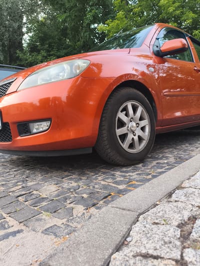 The image shows the front passenger side of an orange car parked on a cobblestone street. The car's headlight, fog light, and a portion of the grille are visible, along with the front wheel and fender. The car appears to be a hatchback, possibly a Mazda. The cobblestone surface is interspersed with sand and small patches of greenery, suggesting an outdoor setting, likely a street or parking area. Trees with lush green foliage form the background, indicating it is likely daytime and possibly spring or summer. The lighting suggests it is not harsh midday sun, but rather a softer light, perhaps late afternoon or early morning. There is no text visible in the image. The setting appears to be a quiet, possibly residential, area in Nowy Dwór Mazowiecki, Poland.