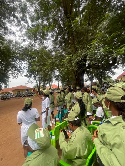 The image depicts an outdoor gathering of people, predominantly young adults, in Emure-Ekiti, Nigeria. They are dressed in matching light green uniforms and caps, suggesting participation in a program or event, possibly National Youth Service Corps (NYSC). The setting appears to be a courtyard or open field, with a dusty red ground surface. Several large trees with dense foliage provide shade over the area, with dappled sunlight filtering through. In the background, there are buildings with orange and yellow facades, indicative of an institutional or educational environment, and a red-roofed structure.

In the foreground and midground, individuals are seen standing and seated in bright green plastic chairs. Many are holding mobile phones, with some actively taking photos or videos. One person in the foreground, from the rear view, is wearing a white t-shirt and white shorts, also in uniform, and is looking towards a group of people gathered further back. Another individual, also viewed from behind, is wearing a white t-shirt and white shorts and is looking to their left.

The group appears to be attentive, possibly listening to an announcement or observing an activity. The overall atmosphere suggests a communal event in progress, under an overcast or partially cloudy sky. The presence of multiple individuals in uniform indicates a structured gathering, possibly related to training or a formal ceremony.