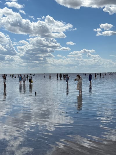 A wide-angle, eye-level shot shows a salt lake with people wading in shallow water. The sky is blue with scattered white clouds. The water is calm and reflects the sky and clouds. A number of people are visible in the distance, scattered across the lake, some walking and some standing. A woman in a light-colored dress stands in the foreground, facing away from the camera. She appears to be holding something in her left hand. In the middle ground, a group of people are gathered, some of them interacting with each other. The overall impression is one of a serene, natural environment being enjoyed by visitors. The location is identified as the Salt Lake in Bursol', Russia.