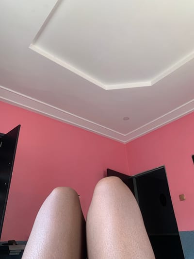 The image shows a low-angle view of a room. The walls are painted a bright pink, and the ceiling is white with decorative molding. In the foreground, the tops of two legs are visible, appearing to belong to a person lying down or sitting. The skin texture on the legs is clearly visible. There are two dark doorways on the right side of the image, one of which is partially open, revealing darkness within. A light switch or electrical outlet is visible on the pink wall near one of the doorways. A black object, possibly a cabinet or furniture, is on the left side of the image. The scene appears to be indoors, likely a residential bedroom or living space. The location is specified as Kakau, Nigeria.