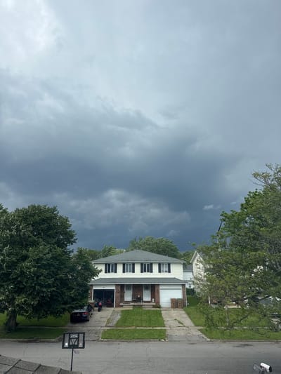 A dark, stormy sky looms over a suburban residential street in the Town of Cheektowaga, United States of America. The image captures a two-story duplex house with a white facade and black trim, featuring a two-car garage. In front of the garage, two trash bins are visible. A black sedan and a motorcycle are parked in one of the driveways. Green trees with lush foliage flank the house, and a basketball hoop stands on the asphalt road. The scene is overcast, with dramatic clouds suggesting an approaching storm.
