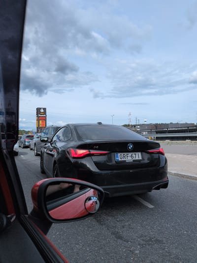 The image shows a line of cars on a wet asphalt road, likely during a traffic jam. The perspective is from inside a red car, looking forward and to the side. A black BMW is prominently featured in the foreground, its rear end facing the viewer. The car has a Finnish license plate that reads "GRF-671". In the background, a tall sign for "ABC Pesukatu" displays gas prices and a thumbs-up icon. Further in the distance, industrial buildings and overhead power lines are visible under a cloudy sky. The scene suggests a location within Helsinki, Finland, possibly near a gas station and car wash. The overall atmosphere is one of urban transit under a brooding sky, with the wet road surface reflecting the ambient light.