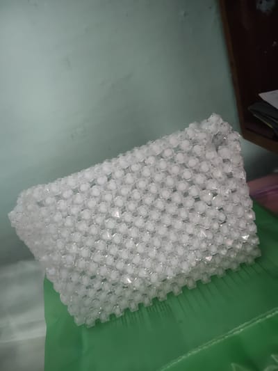 A beaded purse lies on a green surface. The purse is made of translucent white beads, arranged in a honeycomb pattern. The background consists of a light blue wall and a dark wooden shelf. The image appears to be taken indoors, possibly in a home. There are no people visible in the image. The purse is the primary subject, with its intricate beaded construction being a prominent feature.