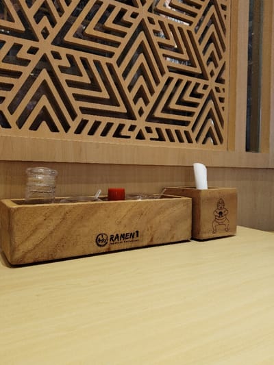The image shows a wooden condiment tray with the logo "Ramen 1 Japanese Restaurant" on the front. The tray is on a wooden table inside a restaurant. There are three small containers on the tray, one of which contains a clear liquid. A small, red lidded container is sitting on the tray as well. A small, wooden container with the image of a cartoon pig on it is sitting on the table to the right of the tray. A white napkin is sticking out of the top of the container. The scene is located at latitude -6.301 and longitude 107.278 in Telukjambe, Indonesia.  The image is taken from a perspective above the table.  The lighting is artificial and there are no people in the image.