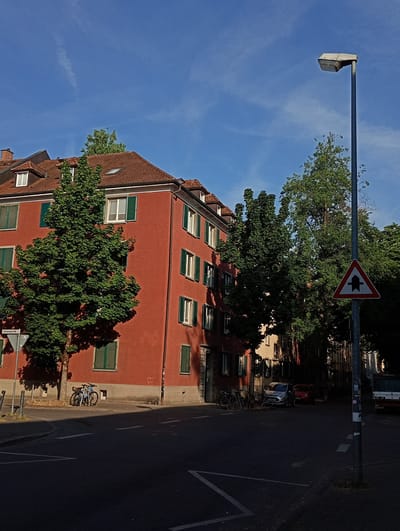 The image depicts a street corner in Constance, Germany, on a clear, sunny day. A large, red, multi-story building with numerous windows and green shutters dominates the left side of the frame. In front of the building, a tree with lush green foliage casts a shadow on the red facade. Two bicycles are parked near the tree.
The street is paved and marked with white lines. A lamppost stands on the right side of the street, with a triangular traffic sign indicating a fork in the road. Behind the lamppost, trees and other buildings are visible. Two cars, one silver and one red, are parked on the street, and further down the road, a bus is partially visible. The sky is a vibrant blue with a few wispy clouds. The overall scene suggests a quiet, residential urban environment bathed in the warm light of a summer day.