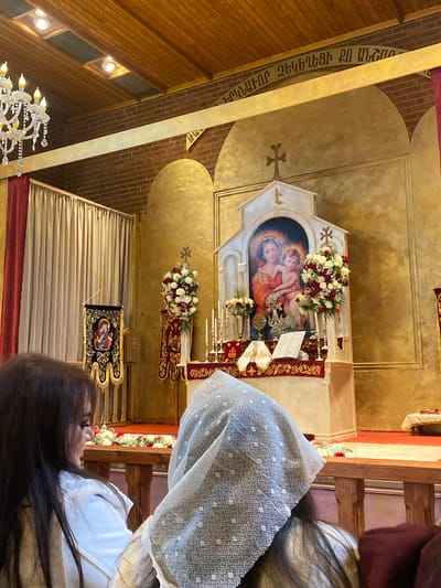 A photograph shows the interior of a church in Södertälje, Sweden. The interior features wooden walls and detailing, including a chandelier. Above the altar are religious artworks depicting the Virgin Mary and Christ child, surrounded by floral arrangements. A woman is visible in the foreground, seated with a white lace head covering. The image is taken from within the church, showing the arrangement of the seating and the altar area. An inscription in Armenian script is visible on a beam in the church.