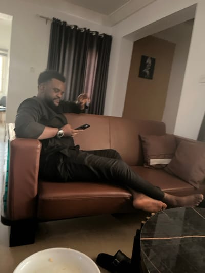 The image displays an indoor scene, likely a living room in Kurudu, Nigeria. A man with a beard is relaxing on a brown, leather-like sofa. He is dressed in a dark long-sleeved top and dark trousers, with a metallic watch on his left wrist.
The man is lying on his back, looking at a smartphone held in his left hand. His right hand appears to be holding a small, dark object, possibly a remote or a snack. His bare feet are visible, resting on the sofa's armrest or backrest on the right side of the frame. Brown cushions are placed on the sofa.
In the foreground, to the right of the sofa, a dark, possibly marbled, round table is partially visible. Below the table, a white bowl containing some food residue is present, along with a dark slipper or sandal on the floor.
The background features dark horizontal blinds or curtains covering a window on the left. Above the curtains, a light-colored wall holds a small, framed black and white picture. The ceiling is also visible, completing the indoor setting. The scene conveys a sense of leisure and comfort. No visible text is discernible in the image.