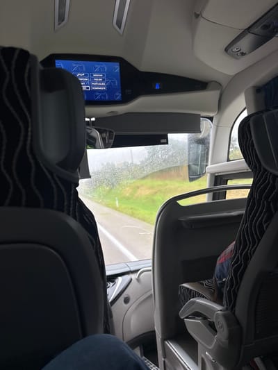 The image depicts the interior of a moving bus in Cantanhede, Portugal, viewed from a seated passenger's perspective looking forward through the front windshield on a rainy day. The windshield is covered with numerous raindrops. Outside, a wet asphalt road with a white solid line is visible, flanked by green grassy areas and distant trees.
Inside, the foreground shows the knees and lower thighs of a person wearing dark blue jeans, indicating the camera's position. The back of the seat directly ahead is dark grey with a wavy pattern on its upper part. To the right, a second passenger is partially visible, occupying a seat of similar design, with a plaid-patterned garment and a bag on the seat. An overhead console is present, featuring a central rectangular display screen showing a language selection menu with options including "MAGYAR," "POLSKI," "PORTUGUÊS," "ROMÂNESTE," "ENGLISH," and an Arabic script option. An illuminated blue seatbelt icon is visible to the right of the display. A small rearview mirror is mounted below the console, and elements of the driver's dashboard are partially visible.