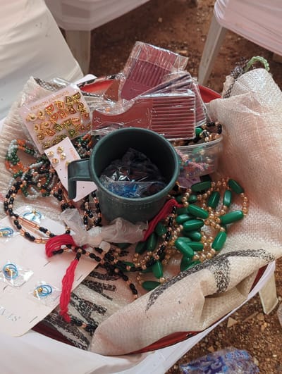 A close-up, overhead view captures a collection of various small goods and accessories contained within a woven bag, likely set in an outdoor or semi-outdoor environment in Unguwar Rimi, Nigeria.
The bag, made of a light-colored, coarsely woven material with dark, patterned sections, is filled with items. Prominently featured are multiple strands of beaded necklaces; one notable type consists of large, oval, green beads interspersed with smaller gold-colored beads. Other necklaces include combinations of black, gold, orange, and multi-colored beads, some tied with a red thread or cord.
To the left, several brown plastic combs are visible, each individually wrapped in clear plastic packaging. A dark green mug sits near the center, also holding some items wrapped in clear plastic. Various earrings are displayed on cardboard backings: one card clearly states "FASHION EARRINGS" and shows numerous small, gold-colored, letter-shaped studs. Another card displays multiple gold stud earrings. Further down, small blue hoop earrings are visible on a card, with the partial word "STA" printed on it, possibly for "Stainless." A small white label with the text "GARANT" and the number "1" is also present, suggesting a guarantee.
The background outside the bag reveals reddish-brown ground, possibly dirt or gravel, and parts of a white fabric structure, which could be a tent or tablecloth. The bright, even lighting suggests it is daytime. No specific activity or individuals are depicted, and no particular emotions are conveyed.