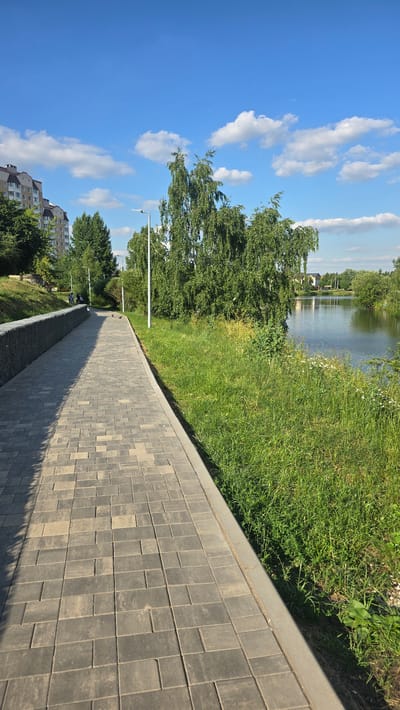 A paved pathway runs alongside a body of water, bordered by lush green grass and trees. The sky is a vibrant blue, dotted with fluffy white clouds, suggesting a pleasant, sunny day. In the distance, across the water, houses and more trees are visible, hinting at a residential area or park. The pathway is constructed of rectangular grey pavers, creating a neat and orderly surface. A gentle slope leads down from the grassy area to the water's edge, where reflections of the sky and trees shimmer on the surface. Further down the pathway, the trees become denser, and the presence of streetlights suggests this is a public walkway designed for both daytime and evening use. In the lower left corner of the frame, the corner of a multi-story building can be seen amongst the trees, indicating the proximity of an urban or suburban environment. The overall scene evokes a sense of peace and tranquility, a perfect spot for a leisurely stroll by the water on a clear day. No visible text is present in the image.