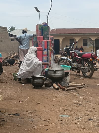 The image depicts an outdoor scene in Tilde, Nigeria, likely during daylight. In the foreground, a person in white clothing is hunched over a cooking pot placed on a fire, with steam rising. This suggests a cooking activity is in progress. Nearby, a stack of colorful rolled mats is propped against a wall, possibly for sale or storage. A motorcycle, predominantly red and black, is parked behind the mats, partially obscuring a person in a light blue shirt and a cap.

The setting appears to be a dusty, informal area, with debris scattered on the ground. In the background, a building with a red, corrugated iron roof and arched windows is visible. Some posters or signs are affixed to the building's exterior. The overall impression is one of everyday life in a community setting, possibly a market or a residential area with commercial activity. There are no explicit indicators of specific emotions or time of day beyond daylight. Some text is visible on posters on the building, but it is not legible in the image.