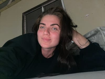 A woman is lying on a bed with her head resting on her arm, looking at the camera. She has long, dark brown hair and is wearing a black sweatshirt. Her skin appears flushed, and she has a small blemish on her chin. In the background, a window with blinds is visible, and a piece of paper is stuck to the wall above it. The room appears to be dimly lit. The scene is likely indoors, possibly in a residential setting.